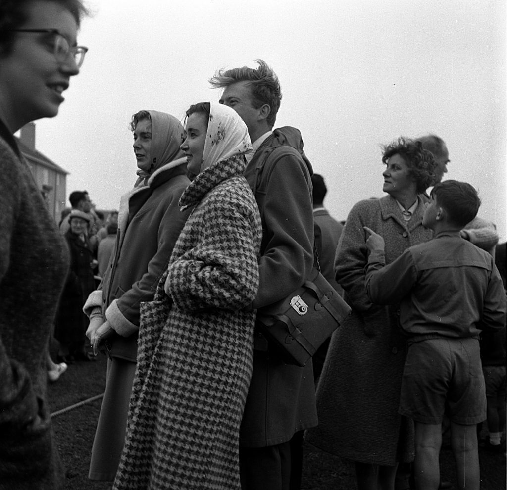 Group of spectators standing outdoors on a grassy field during a sports day event. People are dressed in mid-20th-century clothing, including long coats, patterned overcoats, and headscarves. One individual carries a satchel with a visible clasp. In the background, more people are gathered, and rooftops of buildings can be seen under an overcast sky.
