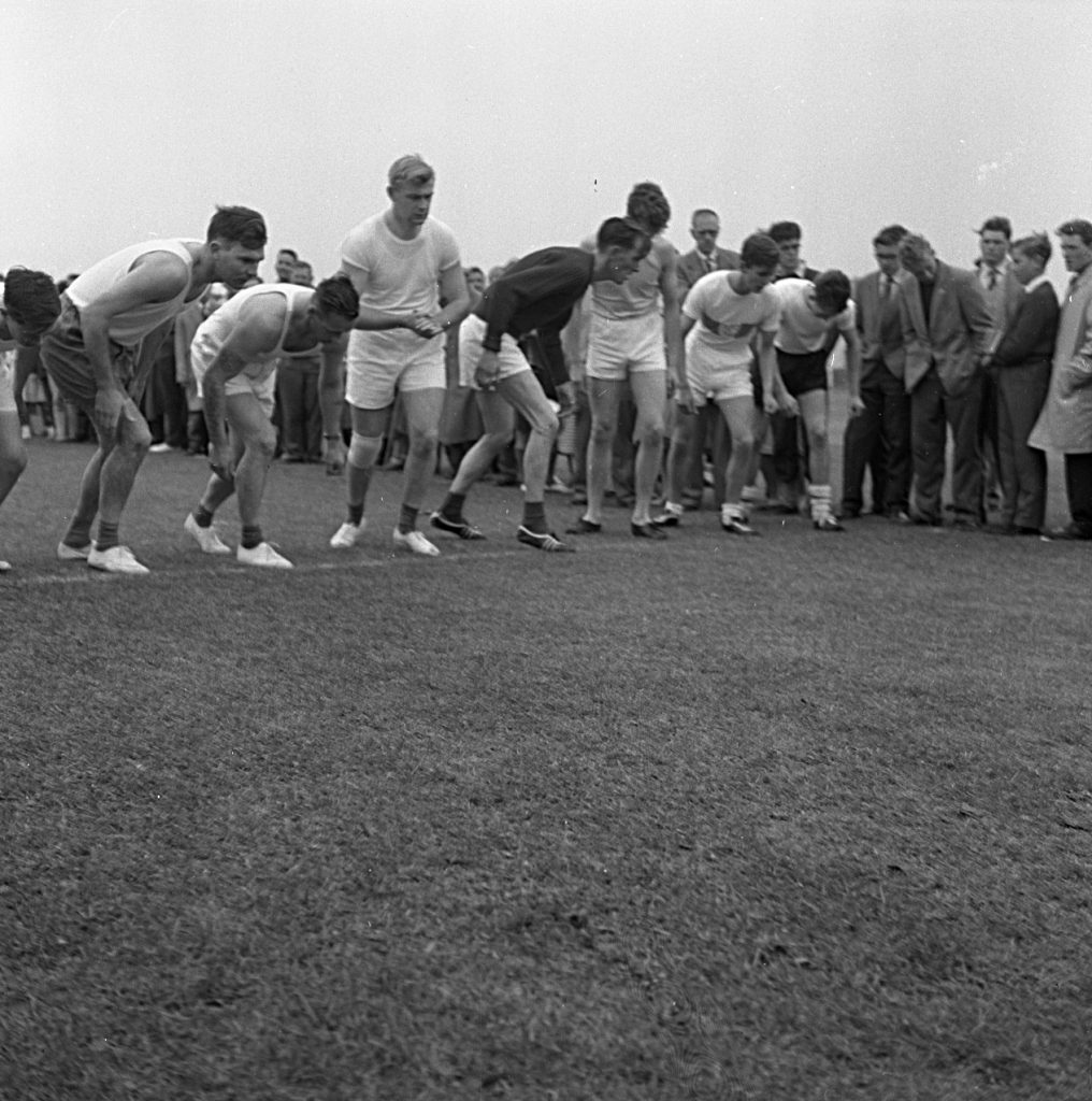 Group of athletes lined up at the starting line on a grassy field during a sports day event. They are wearing athletic clothing, including shorts and light tops, and are bent forward in a ready position. A crowd of spectators stands closely behind them, dressed in mid-20th-century clothing such as suits and coats. The setting is outdoors under an overcast sky.