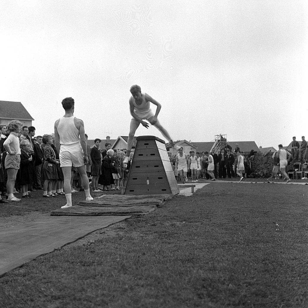An athlete in mid-action climbing over a wooden obstacle during an outdoor sports event on a grassy field. Another athlete stands nearby on a mat, watching. A crowd of spectators lines the left side of the course, while more participants and onlookers are visible in the background near residential buildings.