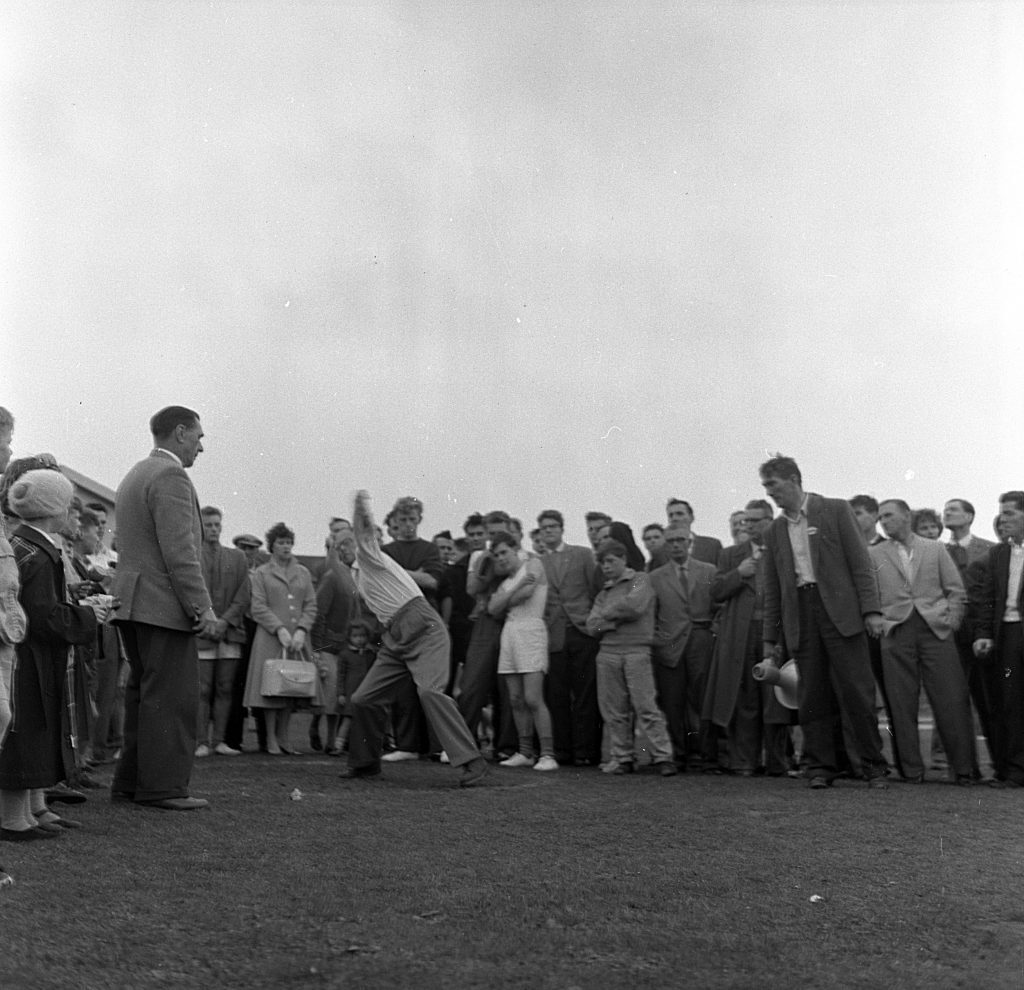A large group of spectators gathered outdoors, watching a person in mid-action throwing an object during a sports event. The thrower is positioned in the center, leaning back with one arm extended behind, preparing to release the object. People of various ages stand closely together, some holding bags, while others observe intently. The scene takes place on a grassy area under an overcast sky.