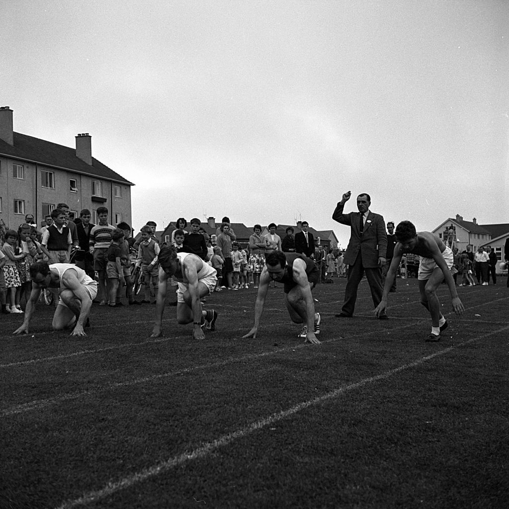 Four athletes crouched in starting positions on a grass track, ready for a race. An official stands behind them with one arm raised, signaling the start. A crowd of spectators, including adults and children, lines the side of the track near residential buildings in the background.