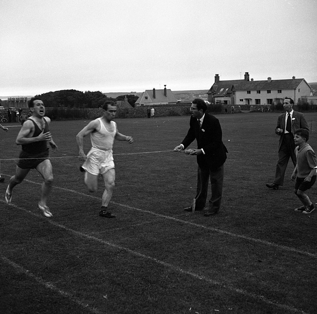 Two athletes running on a grass track during a race, approaching a finish line held by an official in a suit. Several spectators, including a child, stand nearby on the field. In the background, there are houses and bicycles along a fence under an overcast sky.