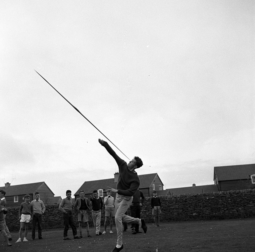 Athlete in mid-action throwing a javelin on a grassy field during a sports day event. The javelin is captured in motion, angled upward against the sky. The thrower is leaning forward with one leg raised for balance, wearing light trousers and a dark sweater. In the background, a group of spectators stands near a stone wall, with houses visible behind them under an overcast sky.