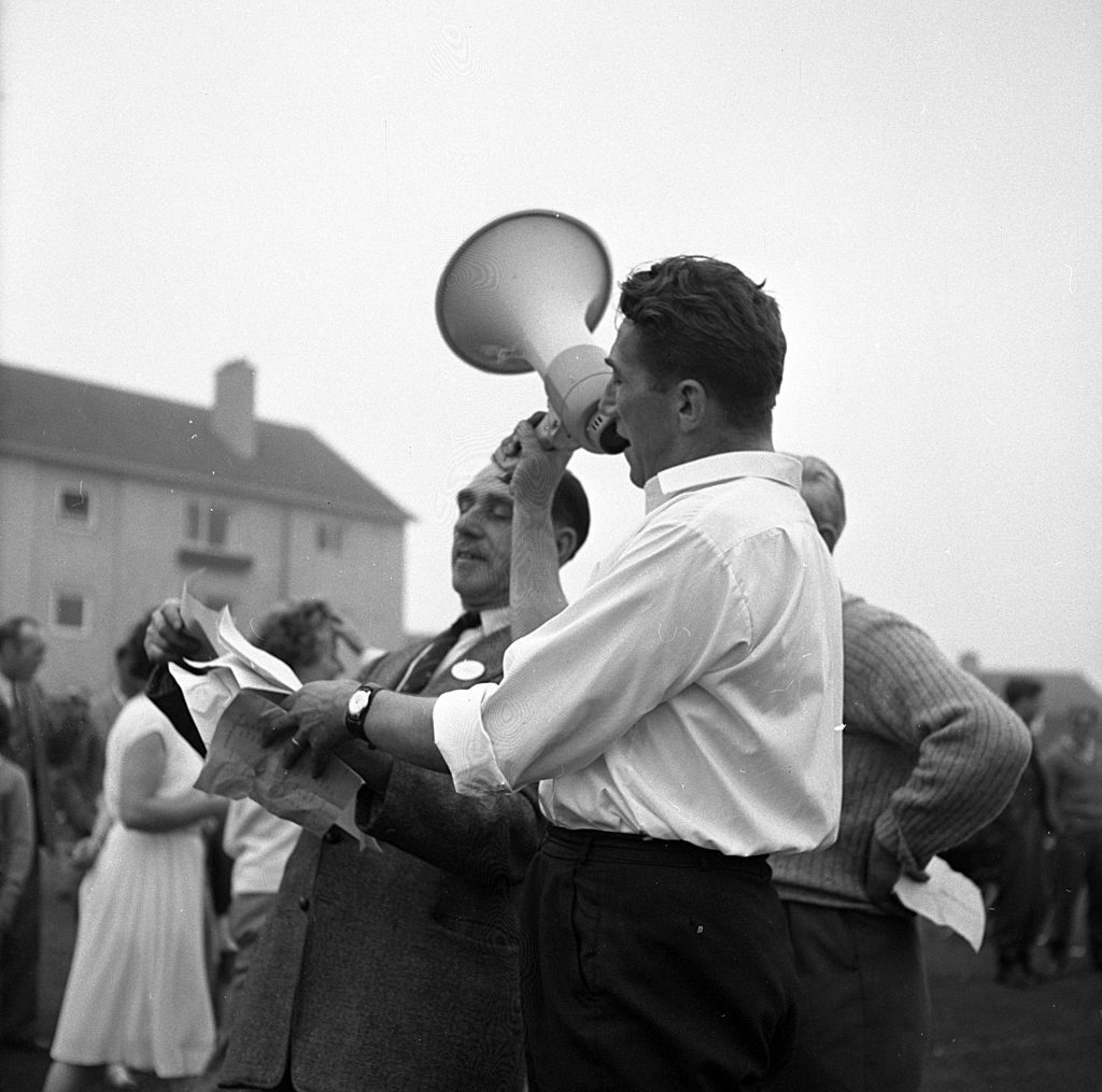 Two individuals standing outdoors during a sports day event, one holding a megaphone and the other holding sheets of paper, appearing to read or announce information. They are dressed in mid-20th-century clothing, including a white shirt and dark trousers. In the background, other people are gathered on a grassy field, with a building visible under an overcast sky.