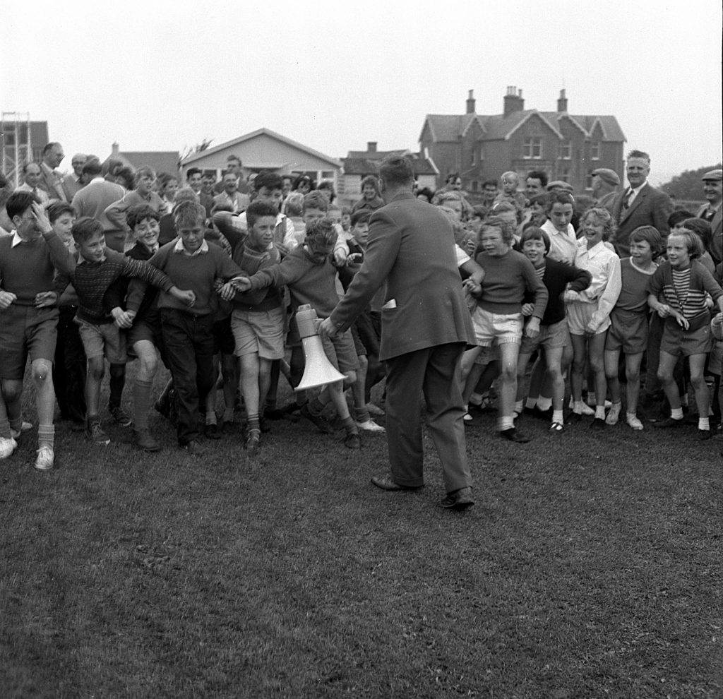 Large group of children lined up on a grassy field at the start of a race during a sports day event. The children are standing shoulder to shoulder, some leaning forward in anticipation. An adult in a suit stands in front of them holding a megaphone, appearing to give instructions or signal the start. Behind the group, a crowd of spectators watches, and several buildings, including houses and a small pavilion, are visible in the background under an overcast sky.