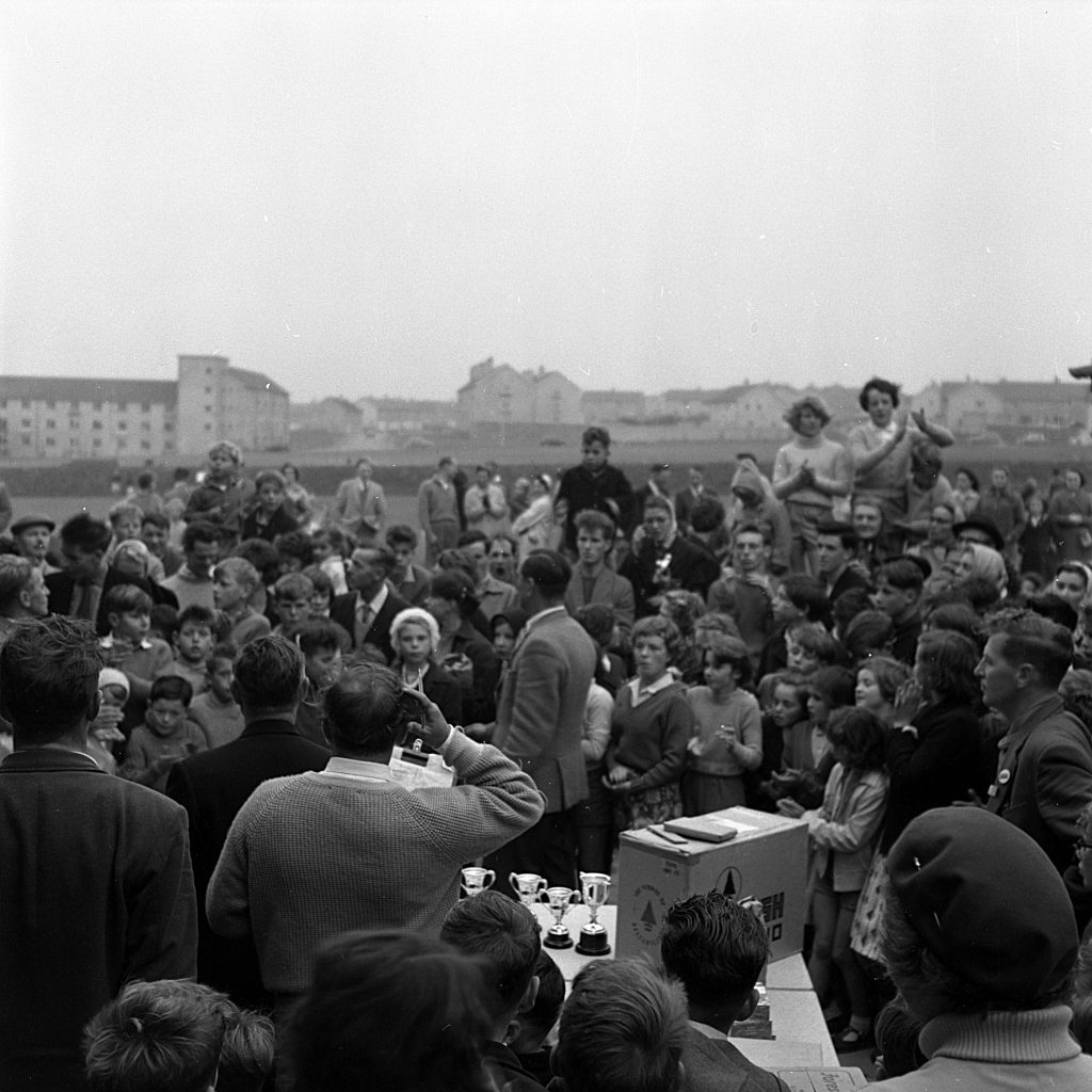 Large crowd gathered outdoors during a sports day event, with people standing closely together around a table displaying several trophies and awards. A man appears to be addressing the crowd near the table, possibly announcing winners or presenting prizes. In the background, more people are visible across the field, and rows of mid-20th-century buildings can be seen under an overcast sky.
