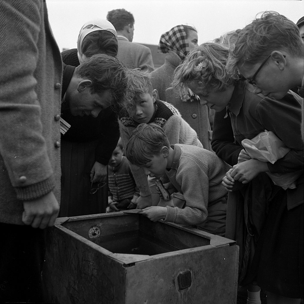 Group of children and adults gathered closely around a wooden box on the ground during an outdoor event. Several children are leaning forward, peering into the box with curiosity, while others stand nearby observing. The box appears to contain water or an object of interest. One child peering into the box appears is poised to throw a coin into it. People are dressed in mid-20th-century clothing, and the setting is a grassy area under an overcast sky.