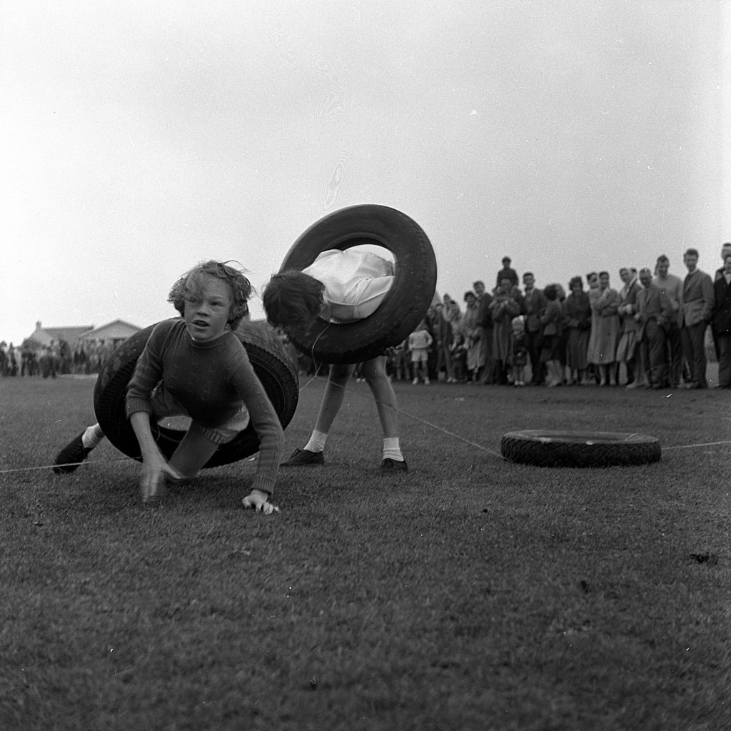 Two children participating in a race involving car tyres on a grassy sports field during a community sports day. One child is crawling forward through a large rubber car tyre, while the other is standing and maneuvering with the ring around the torso. A crowd of spectators stands in the background, watching the event. The setting is outdoors under an overcast sky.