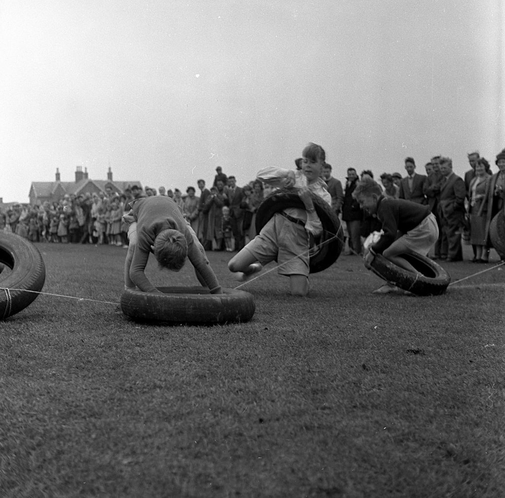 Children participating in a race involving car tyres on a grassy sports field during a community sports day. Each child is maneuvering through or wearing a large rubber ring while crawling or running along marked lanes. A crowd of spectators stands in the background, watching the event. Houses and buildings are visible in the distance under an overcast sky.