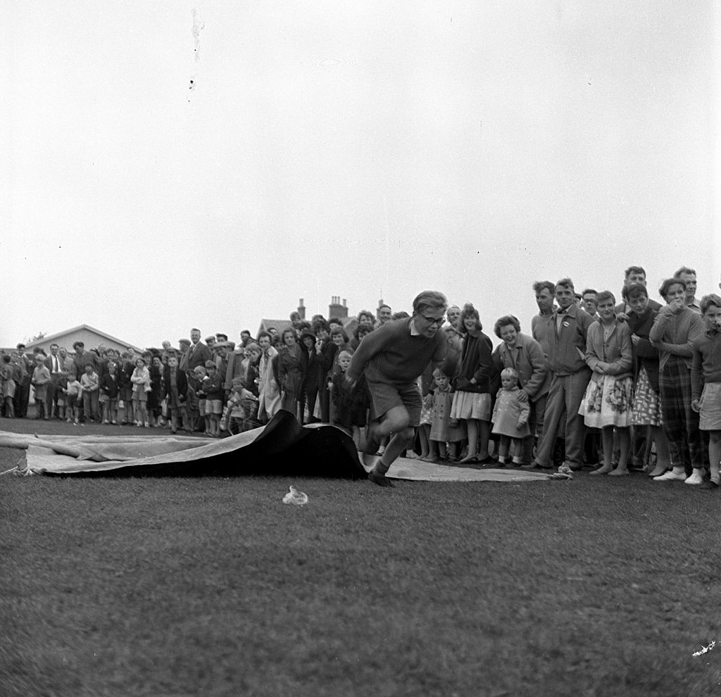 Child running across a grassy field during a sports day event, emerging from a mat he has just crawled under as part of a race. A large crowd of spectators, including adults and children, stands closely behind the marked area, watching the activity. In the background, small buildings and houses are visible under an overcast sky.