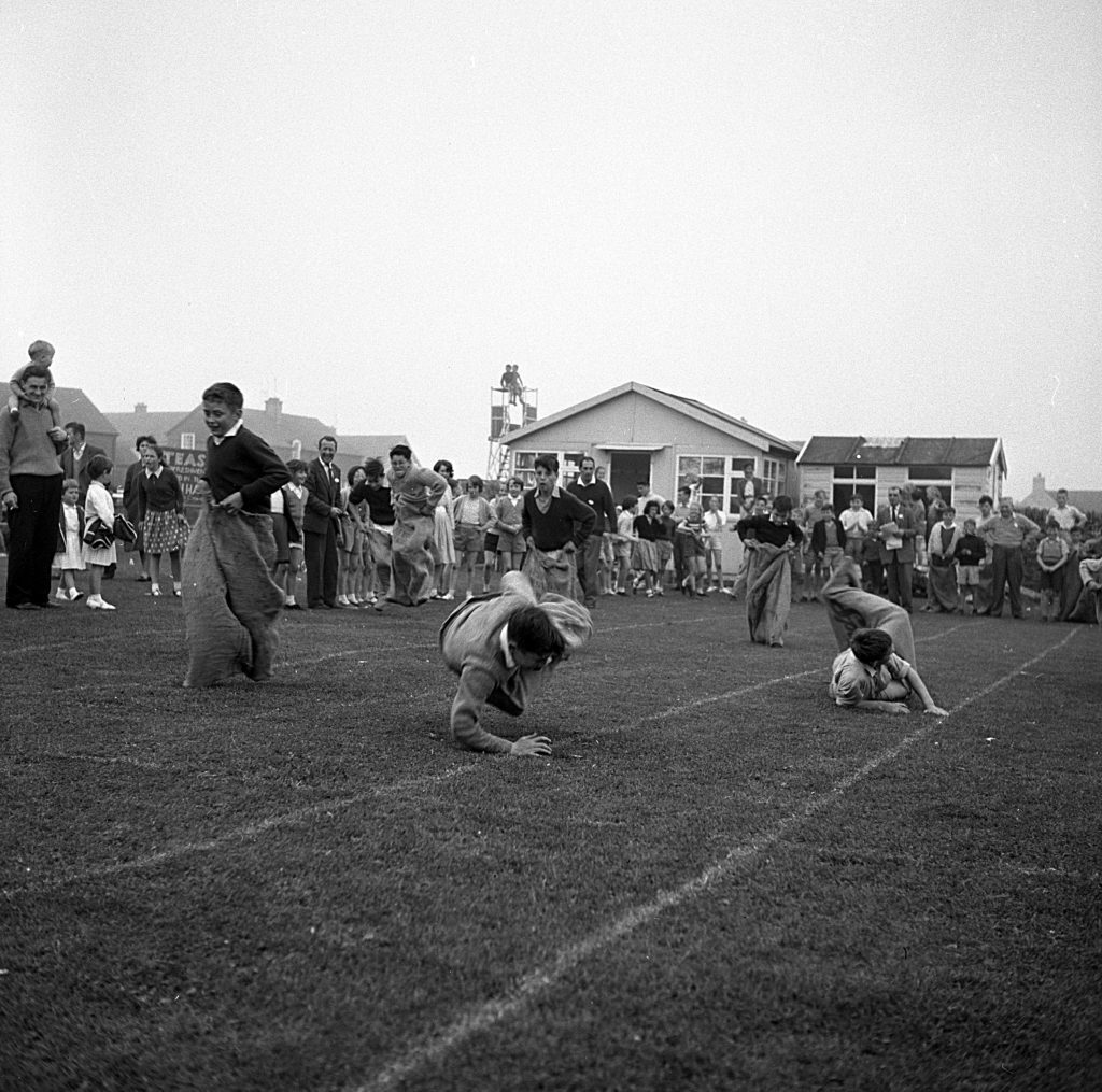 Children participating in a sack race on a grassy sports field during a community sports day event. Several children are inside large sacks, hopping or crawling forward along marked lanes. A crowd of spectators, including adults and children, stands in the background watching. Behind the crowd, there are small buildings and houses, and the sky appears overcast.