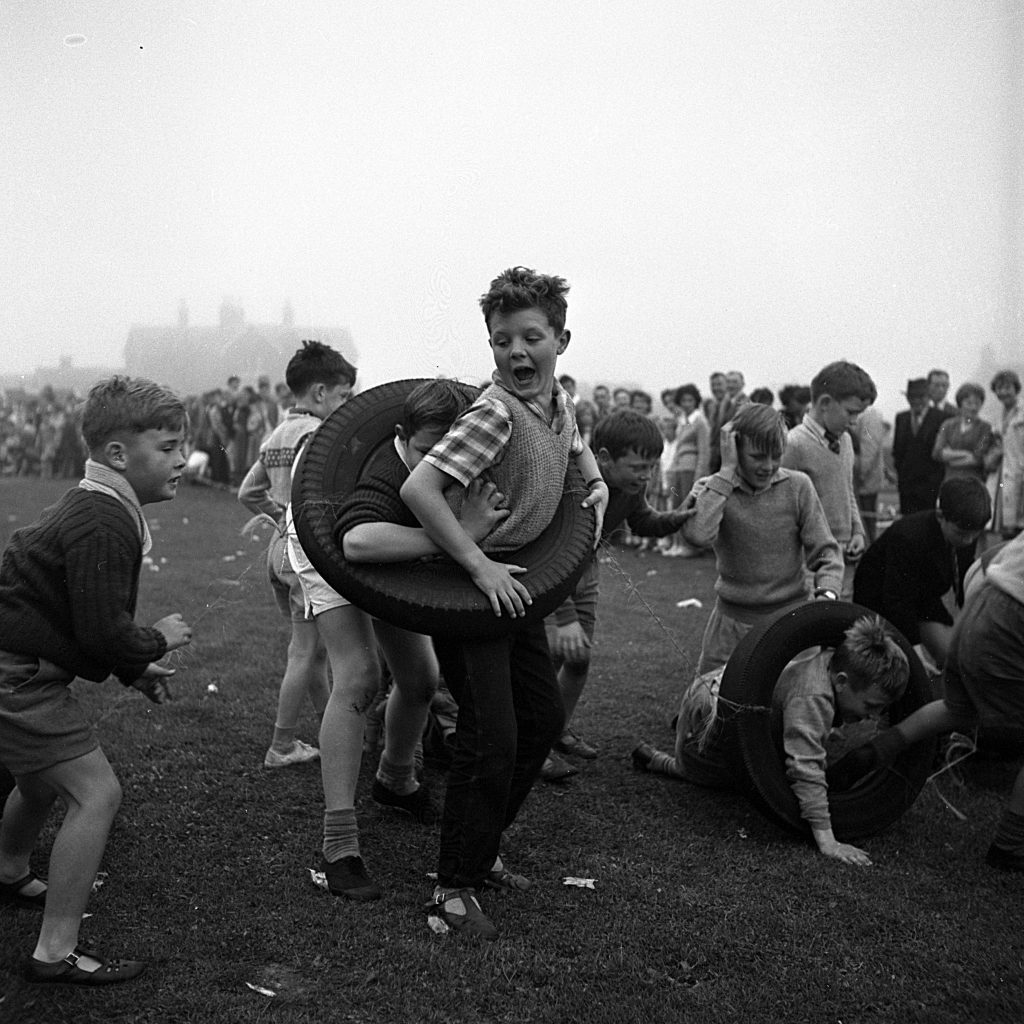 Children taking part in a playful race involving car inner tubes on a grassy field during a sports day event. Some children are wearing the large rubber tyres around their bodies while running, while others are crawling or struggling to move through them. A crowd of spectators stands in the background watching the activity. The setting is outdoors on an overcast day, with houses faintly visible in the distance.