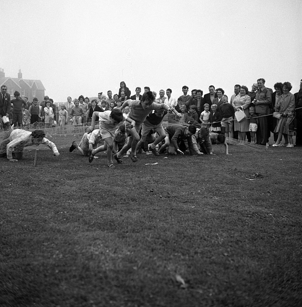 Children participating in a crawling race on a grassy field during a sports day event. Several children are moving forward on hands and knees within marked lanes, while a large crowd of spectators stands in the background watching. The setting appears outdoors on an overcast day, with houses visible in the distance.