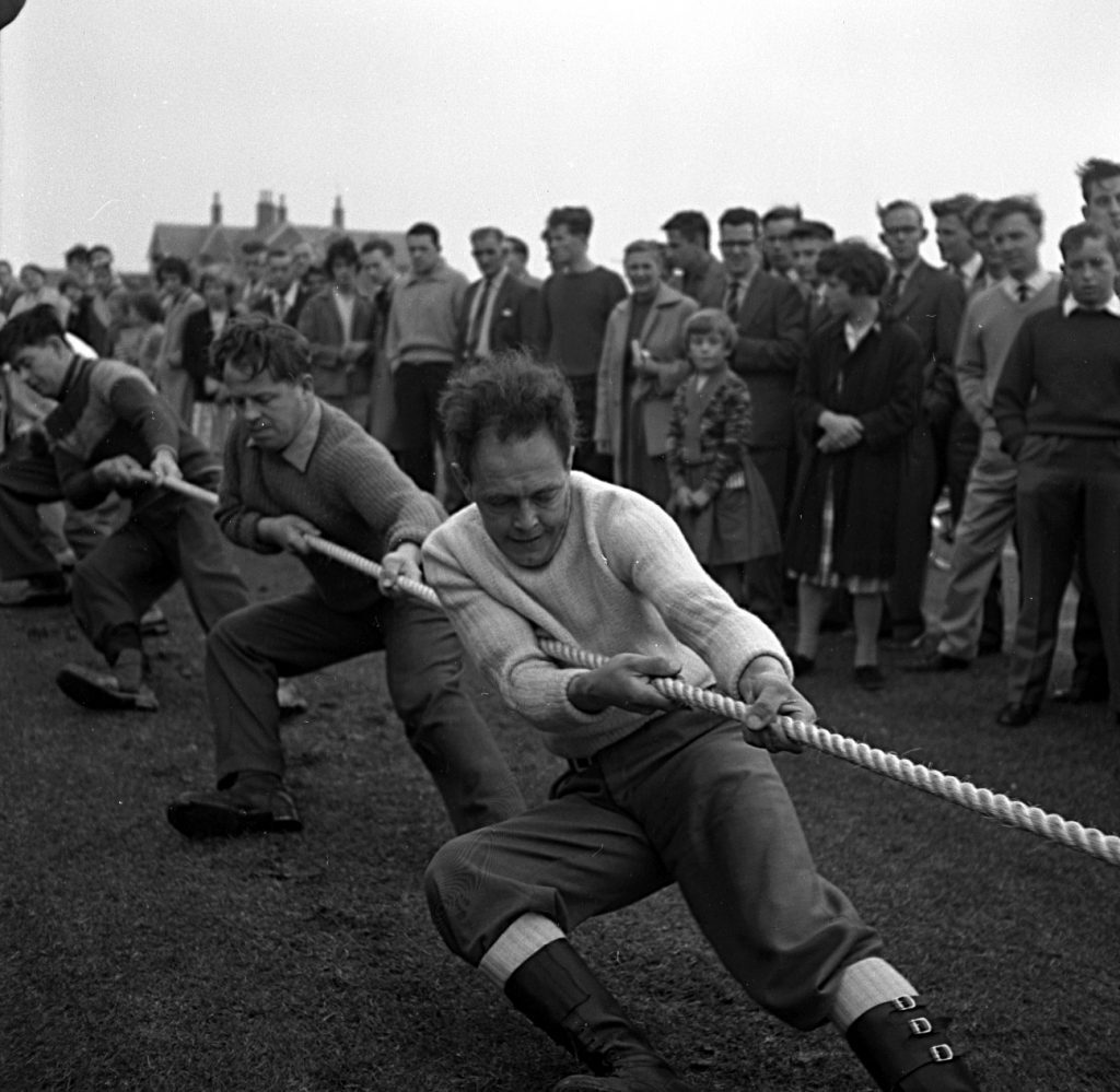 Tug-of-war competition on a grassy field during a sports day event. Several participants are pulling a thick rope with intense effort, leaning backward for leverage. They are dressed in mid-20th-century clothing, including sweaters and boots. Behind them, a large group of spectators stands closely watching, forming a line across the background. The setting appears outdoors with overcast skies and buildings visible in the distance.