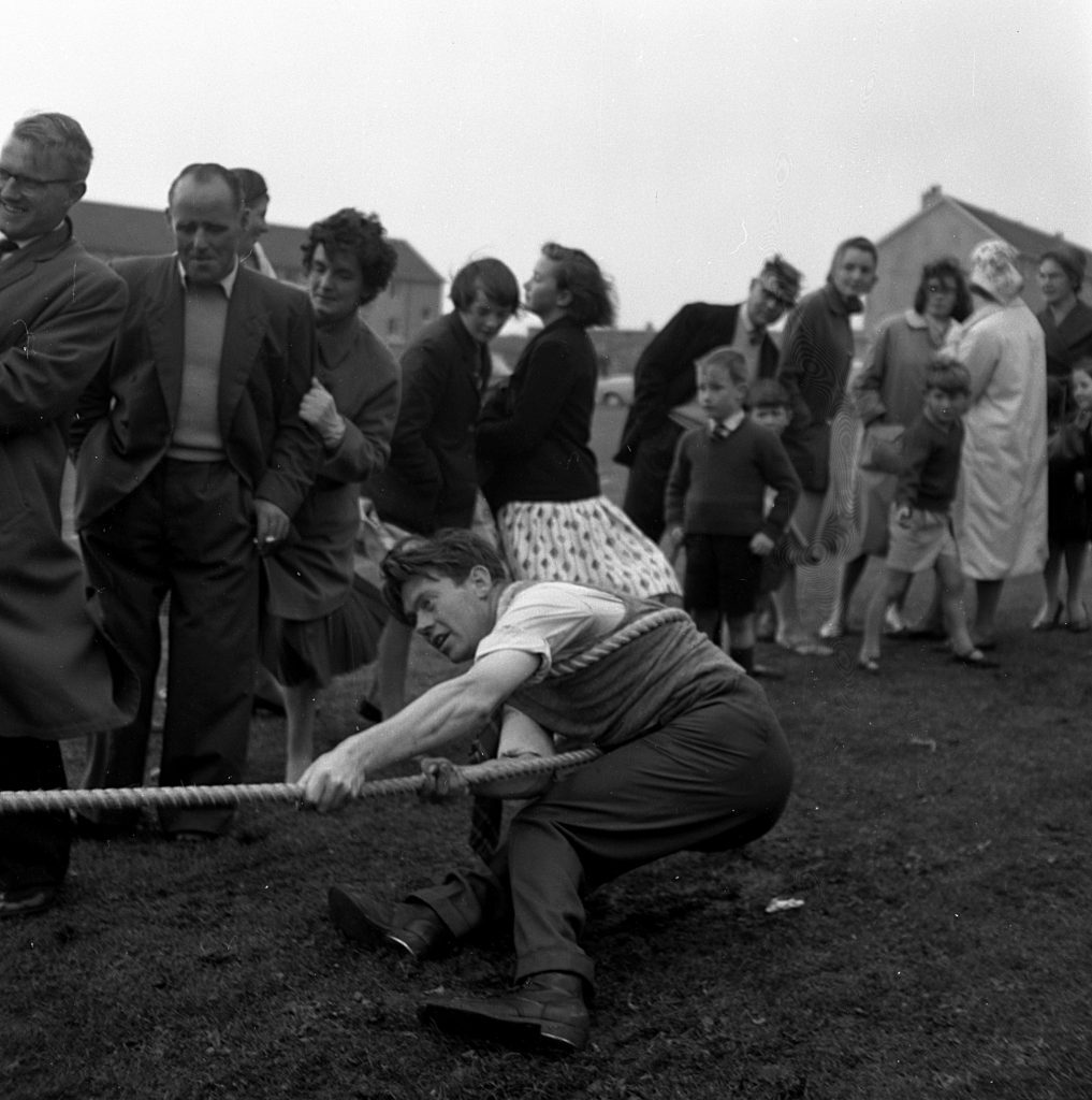 Outdoor tug-of-war competition on a grassy field during a sports day event. A participant in the foreground is crouched low, pulling hard on a thick rope with both hands, leaning back for leverage. Behind, a group of spectators stands in a line watching, including adults and children dressed in mid-20th-century clothing. In the background, houses are visible under an overcast sky.