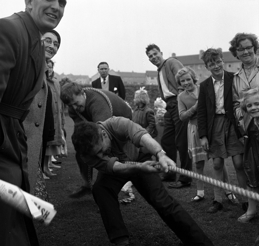 Outdoor scene of a tug-of-war competition on a grassy field during a sports day event. A man in the foreground is pulling hard on a thick rope, leaning backward with effort. Another participant is visible behind him, also gripping the rope. Spectators, including adults and children, stand nearby watching the contest. The background shows rows of houses and an overcast sky.