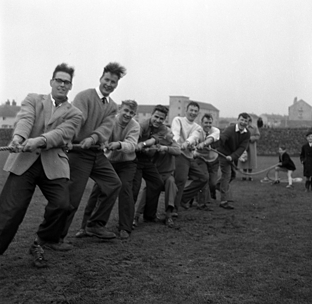 A tug-of-war team pulling a thick rope during an outdoor sports day event on a grassy field. The group consists of several men positioned in a line, leaning back with effort as they pull. They are dressed in mid-20th-century clothing, including jackets, sweaters, and trousers. In the background, spectators stand watching, and rows of buildings are visible under an overcast sky.