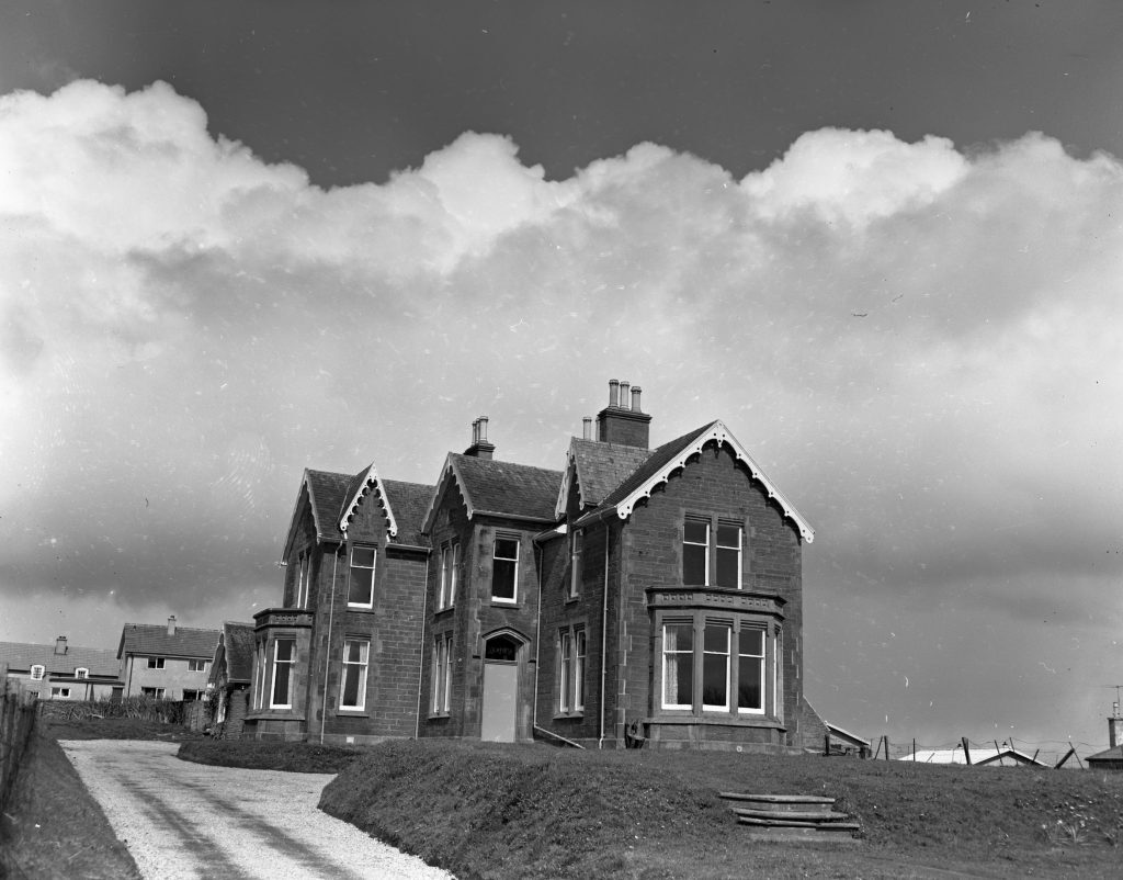 Black and white photograph of a large two-storey stone house with steep gabled roofs and decorative bargeboards. The building has multiple chimneys and tall windows, with a central doorway. A gravel driveway leads up to the house, and steps are visible on the grassy slope in the foreground. Additional houses can be seen in the background under a sky with large, billowing clouds.