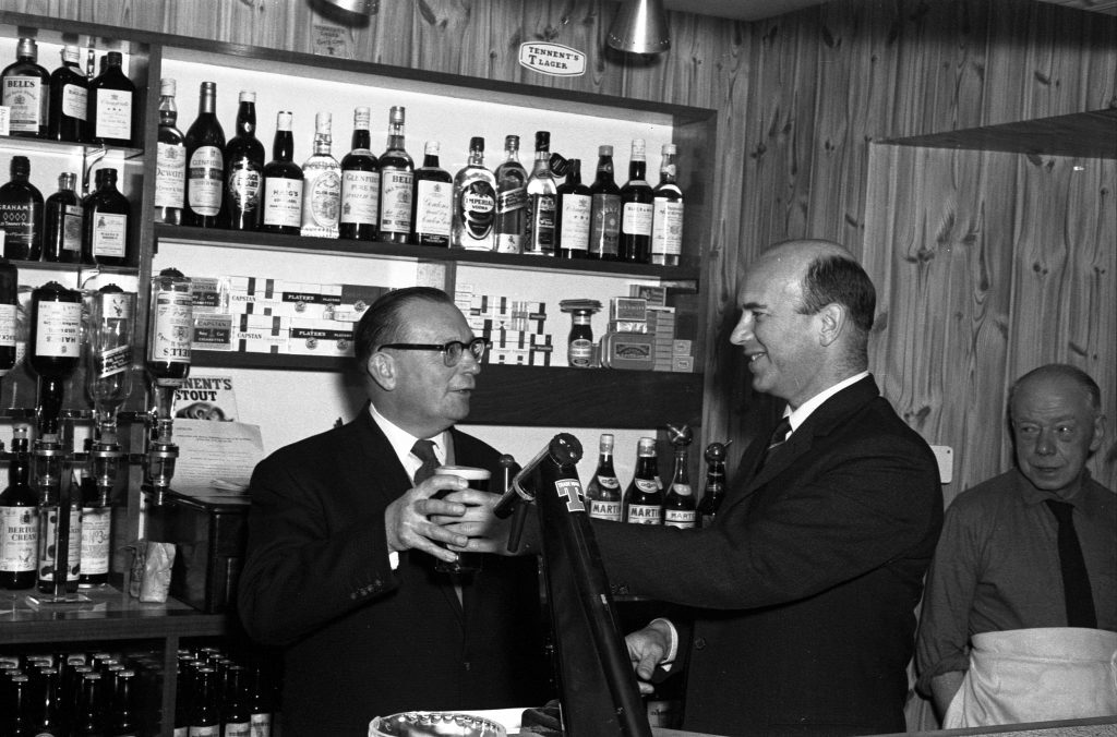 Two individuals standing behind a bar counter in a wood-paneled room. One person is holding a glass near a beer tap, while the other appears to be assisting. Shelves behind the counter are stocked with bottles of alcohol and boxes, and a sign reading ‘Tennent’s Lager’ is visible above the shelves.