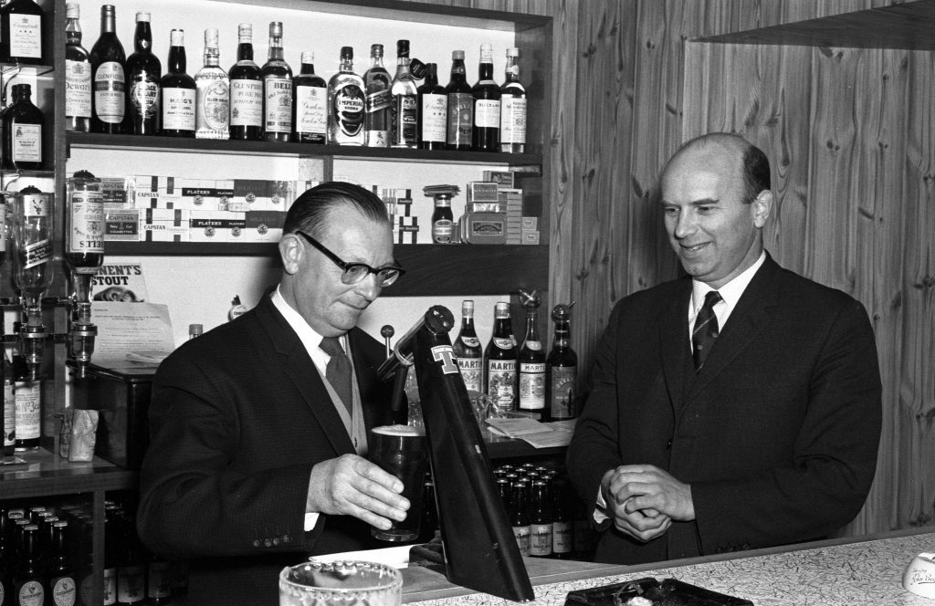 Two individuals standing behind a bar counter in a wood-paneled room. One person is operating a beer tap, while the other stands nearby with hands clasped. Shelves behind the counter are stocked with various bottles of alcohol and boxes, and the countertop has a tray and a glass.