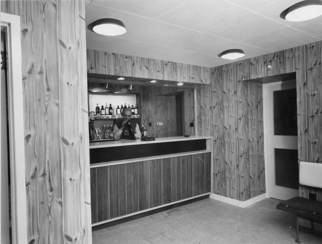 Interior view of a small bar area with wood-paneled walls and ceiling. The bar counter is made of matching wood with a dark strip across the front. Behind the counter, shelves display bottles of alcohol and a few promotional items. The space is lit by round ceiling lights and recessed lights above the bar. A doorway and a bench are visible to the right.
