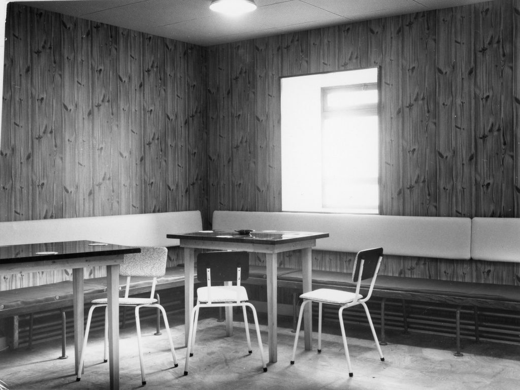 Interior of a small lounge or seating area with wood-paneled walls and a single window. The room contains two rectangular tables with simple chairs around them and a long cushioned bench running along the walls.