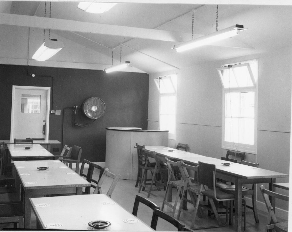 Interior of a club room with several rectangular tables arranged in rows, surrounded by wooden chairs. The room has large windows on the left, fluorescent lights hanging from the ceiling, and a bar is in the far right corner. Ashtrays and coasters are placed on the tables, and the floor appears to be linoleum.”