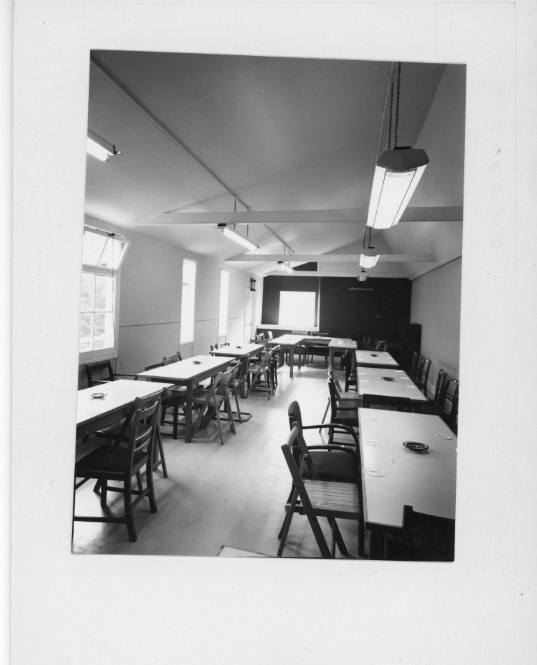 Interior of a club room with several rectangular tables arranged in rows, surrounded by wooden chairs. The room has large windows on the left and fluorescent lights hanging from the ceiling. Ashtrays and coasters are placed on the tables, and the floor appears to be linoleum.