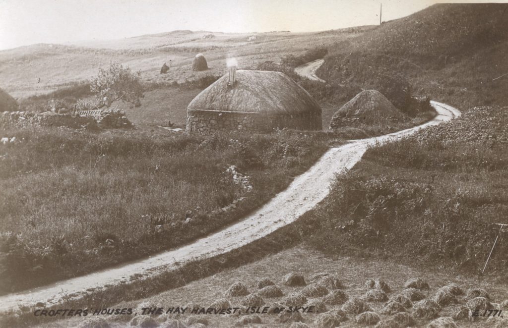 A black-and-white photograph of a rural landscape on the Isle of Skye, showing traditional crofters’ houses with rounded stone walls and thatched roofs. A winding dirt road leads through grassy fields toward the houses, with haystacks visible nearby. The foreground features cut hay spread across the ground, and rolling hills stretch into the background under an open sky.