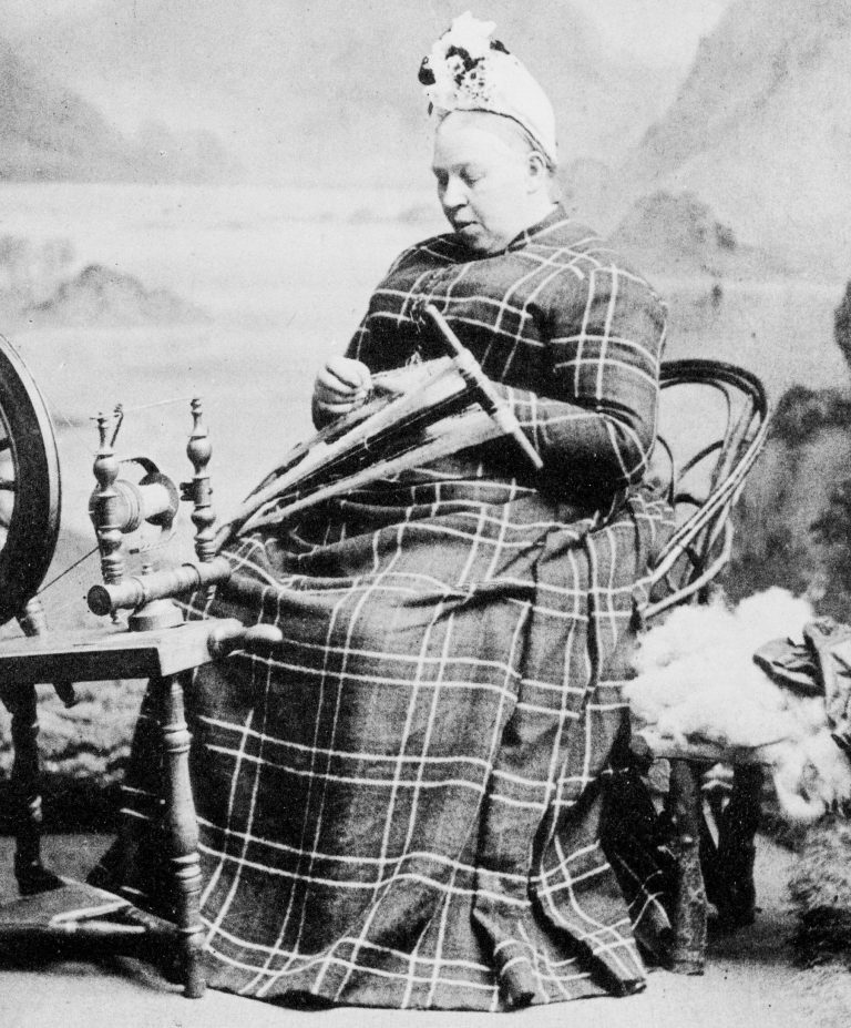 Black-and-white photograph showing a woman seated on a wooden chair, wearing a long, plaid-patterned dress and a head covering. The individual is holding a traditional spinning tool, with a spinning wheel positioned to the left. To the right, there is a pile of raw wool on another chair. The background features a painted scenic backdrop, suggesting a studio setting.