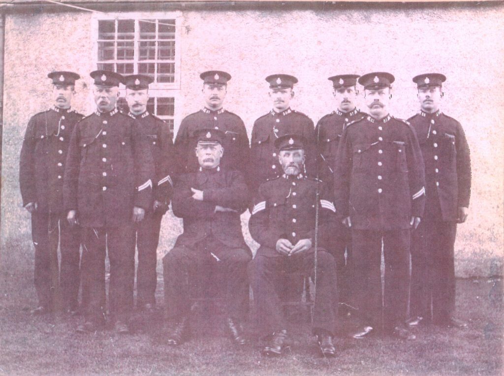 Photograph showing a group of uniformed police officers posed in two rows outside a building. The officers are wearing dark formal uniforms with high collars, metal buttons, and peaked caps. Some uniforms display chevrons on the sleeves, indicating rank. Two officers are seated in the front row, while the others stand behind them. The background features a rough-textured wall with a multi-paned window, and the group is standing on grass.