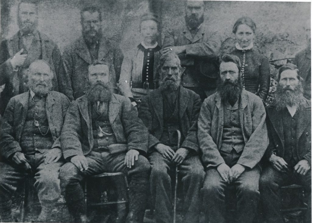 black-and-white group photograph showing eight men and two women seated and standing in two rows. The individuals are dressed in formal, heavy clothing typical of the late 19th or early 20th century, including jackets, waistcoats, and high-collared shirts. Some have visible long beards, and the seated figures rest their hands on their knees. The background appears to be a plain textured wall or outdoor stone surface