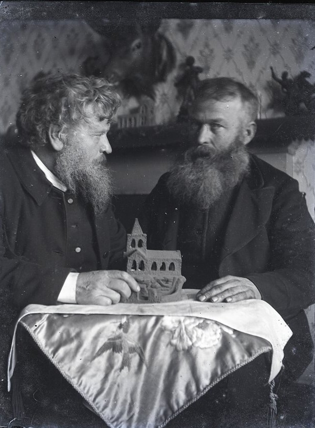 Black and white image of two men sitting down at table, holding a wooden sculpture of a church