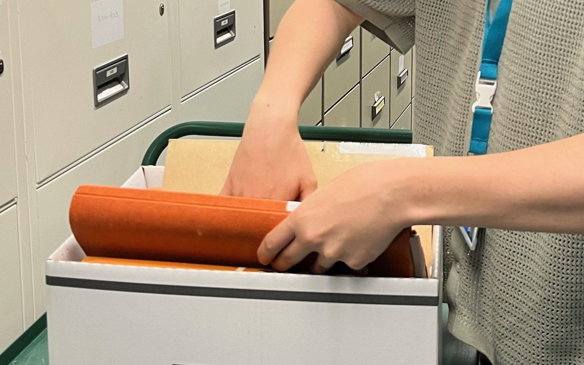 A person's hands can be seen taking a volume from a cardboard storage box