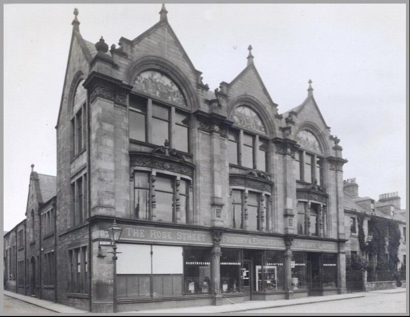 Historic black-and-white photograph of a large stone building with ornate architectural details, including three prominent gables with decorative carvings and large arched windows. The ground floor has signage reading “The Rose Street Foundry & Engineering Company Limited,” with shopfront windows displaying goods. A streetlamp stands at the corner, and the building is situated on a quiet street with adjoining structures.