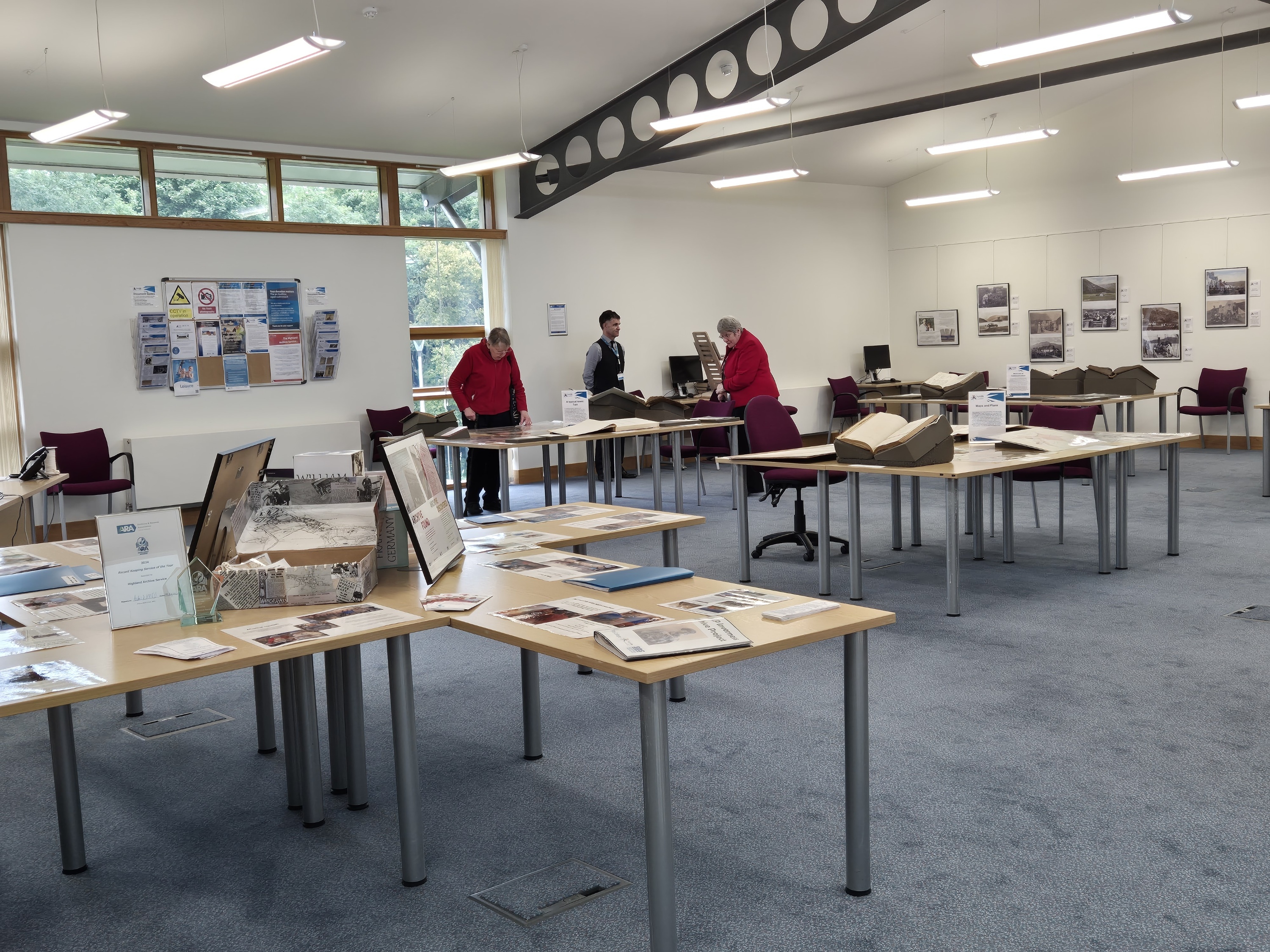 Archive searchroom with exhibition displays and historic documents laid out on tables for visitors to explore. Large archival volumes, photographs, information panels and local history materials are arranged throughout the bright room, with a small number of visitors viewing the displays. Framed historic photographs hang on the walls, and large windows provide natural light.