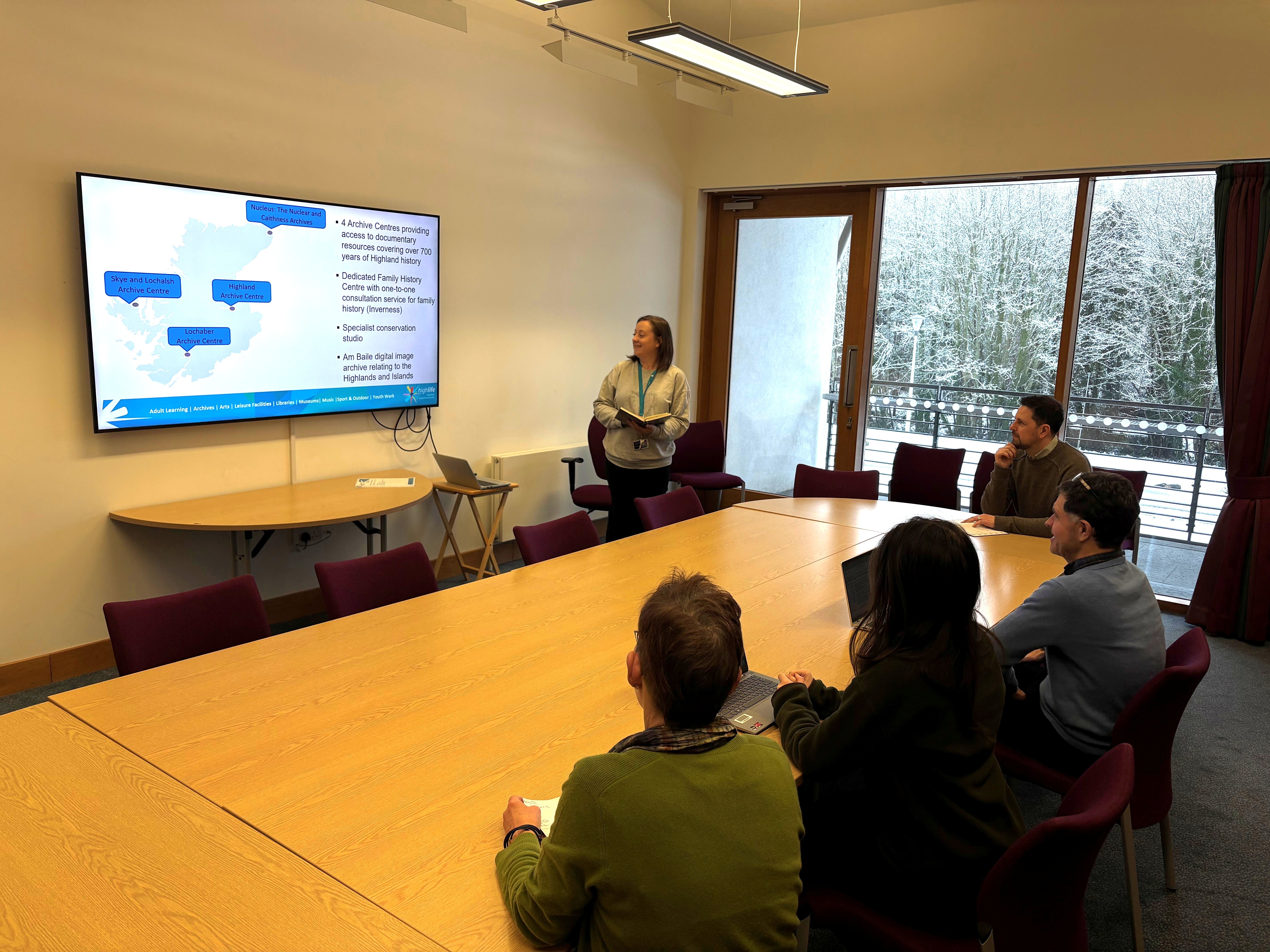 A group of four people seated around a large wooden conference table in a meeting room, watching a presentation displayed on a wall-mounted screen. The screen shows a map with labeled blue boxes and text. One person is standing near the screen, holding a laptop and presenting. The room has large windows in the background. The chairs around the table are upholstered in a dark red color, and the lighting is bright with ceiling fixtures.