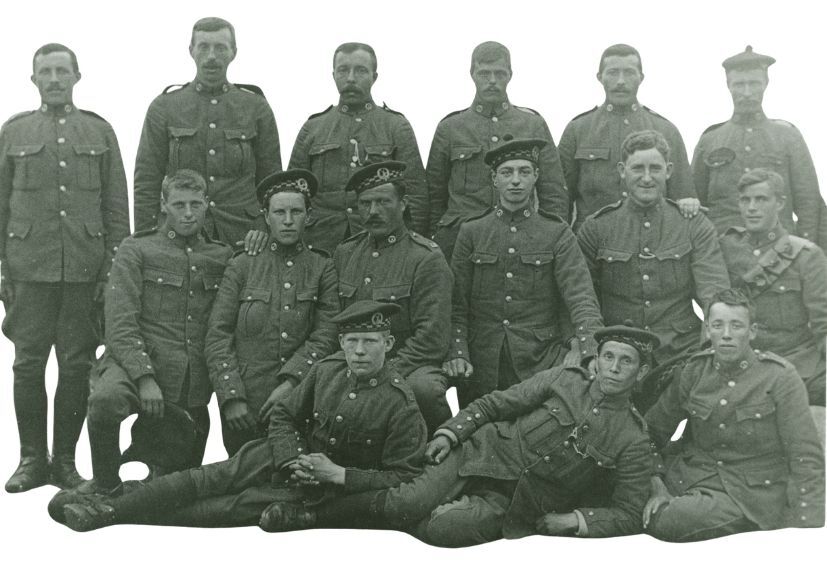 Black-and-white First World War era group photograph showing fourteen uniformed soldiers posed in three rows. Eight soldiers stand at the back, three are seated in the middle, and three recline at the front. The image is cropped to remove the background, focusing attention on the soldiers and their military uniforms.