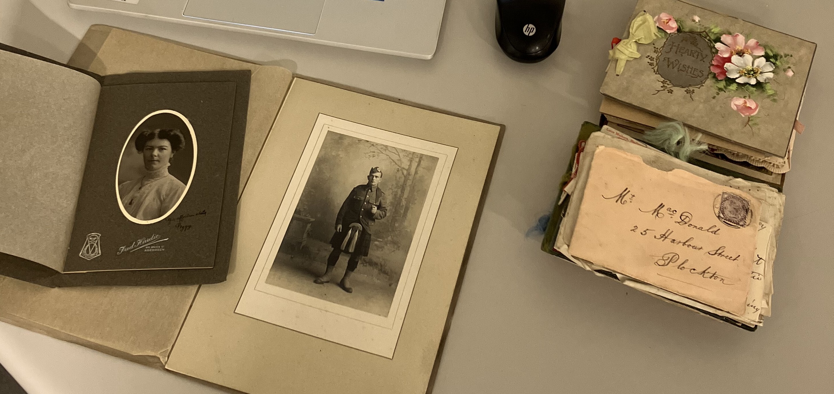 Historic photographs and family correspondence laid out on a table, including a vintage portrait album, a mounted studio photograph of a person in military uniform, and a stack of handwritten letters and greeting cards, illustrating archival materials being preserved and catalogued.