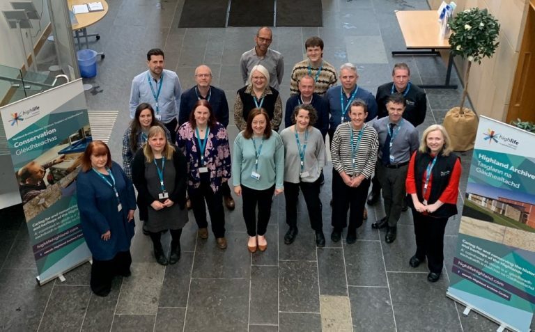 Group photo of staff standing indoors on a stone-tiled floor between two vertical banners. The left banner reads “Conservation Glèidheadhchas” with images of archival materials and the High Life Highland logo. The right banner reads “Highland Archive Taghlaran na Gàidhealtachd” with images of archive facilities and the same logo.