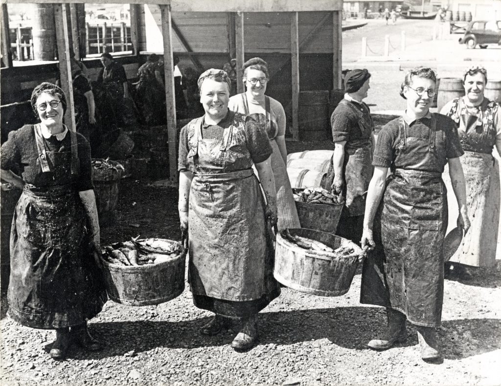 Historic black-and-white photograph showing a group of female workers outdoors at a fish processing site. Several individuals are wearing heavy aprons and head coverings, carrying large metal tubs filled with fish. In the background, there are wooden structures, barrels, and additional workers engaged in similar tasks. The setting appears to be an early 20th-century herring industry scene.