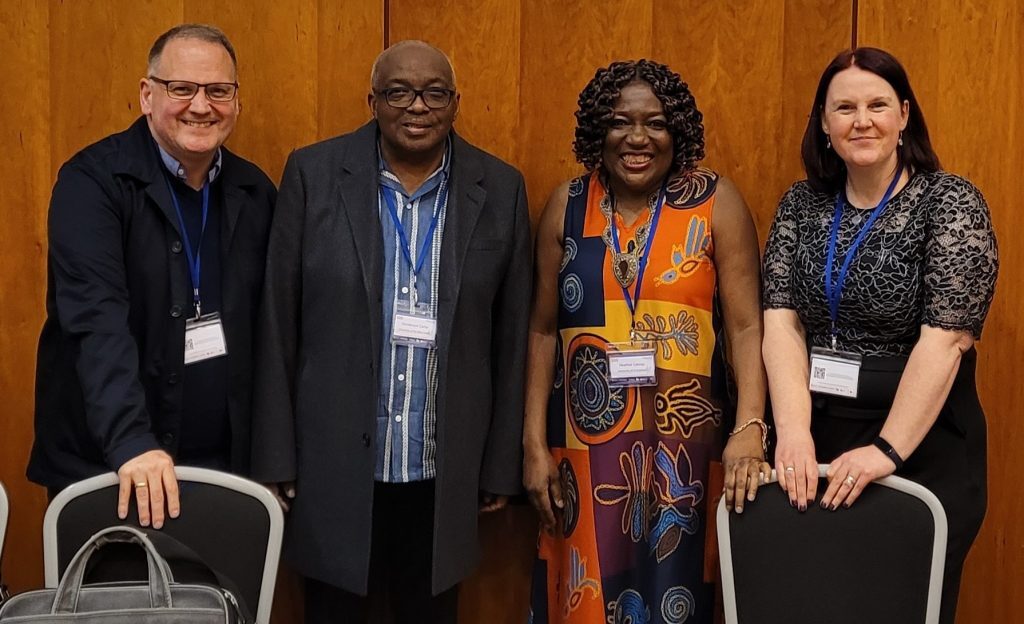 Four individuals standing indoors in front of a wooden-paneled wall, wearing conference name badges and blue lanyards. Two chairs are positioned in front of them, with a bag resting on one chair. The individuals are dressed in formal and semi-formal attire, including a patterned dress, a lace top, a collared shirt with a coat, and a dark jacket. The setting appears to be a professional or academic event.