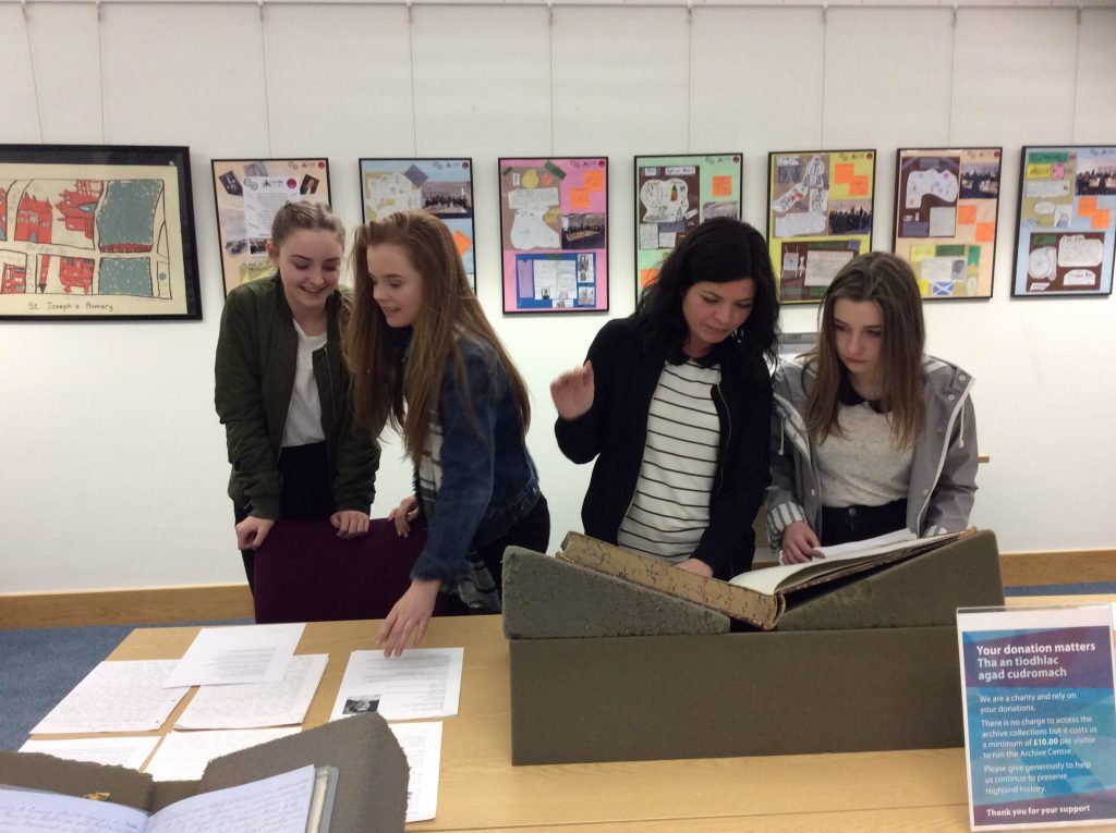 A group of four people standing around a display table in an exhibition space, examining large archival books and documents. The table holds open volumes in protective boxes and several loose sheets of paper. Behind them, a wall features framed maps and colorful posters arranged in a row.