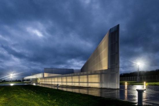A modern, angular building with sleek metallic panels and illuminated sections, photographed at dusk under a dramatic, cloudy sky. The structure features sharp lines and a prominent vertical element on the right side, with warm lighting highlighting its architectural design. The foreground includes a wet pavement reflecting light and a grassy area, while outdoor lamps provide additional illumination around the building.