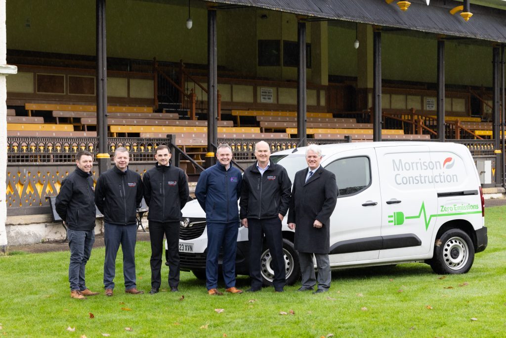 Six adult males representing Morrison Construction and The Highland Council are standing in front of the Northern Meeting Park pavilion and a white Morrisons construction van.