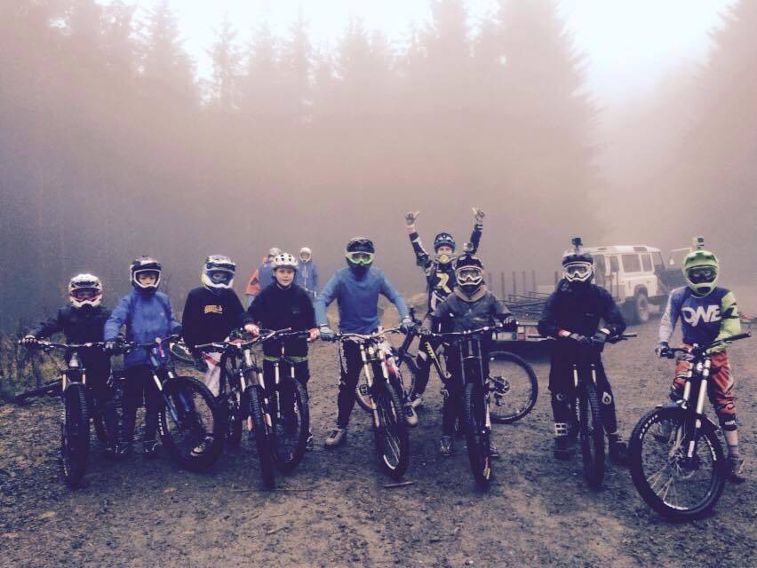 Nine young people on standing with mountain bikes. They are all wearing bike helmets and some are wearing goggles.