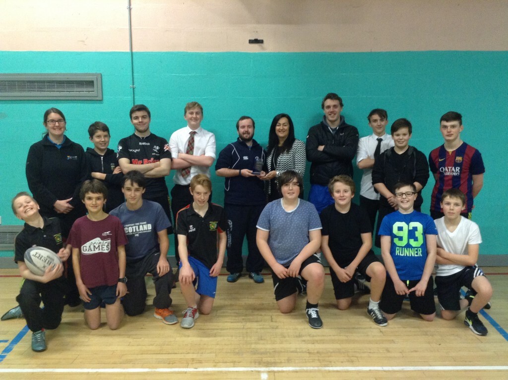 A group of 18 people - both adults and high school students in a gym hall setting. Eight people are in the front row kneeling with one boy holding a rugby ball, The  rest are standing behind. They are all smiling at the camera.