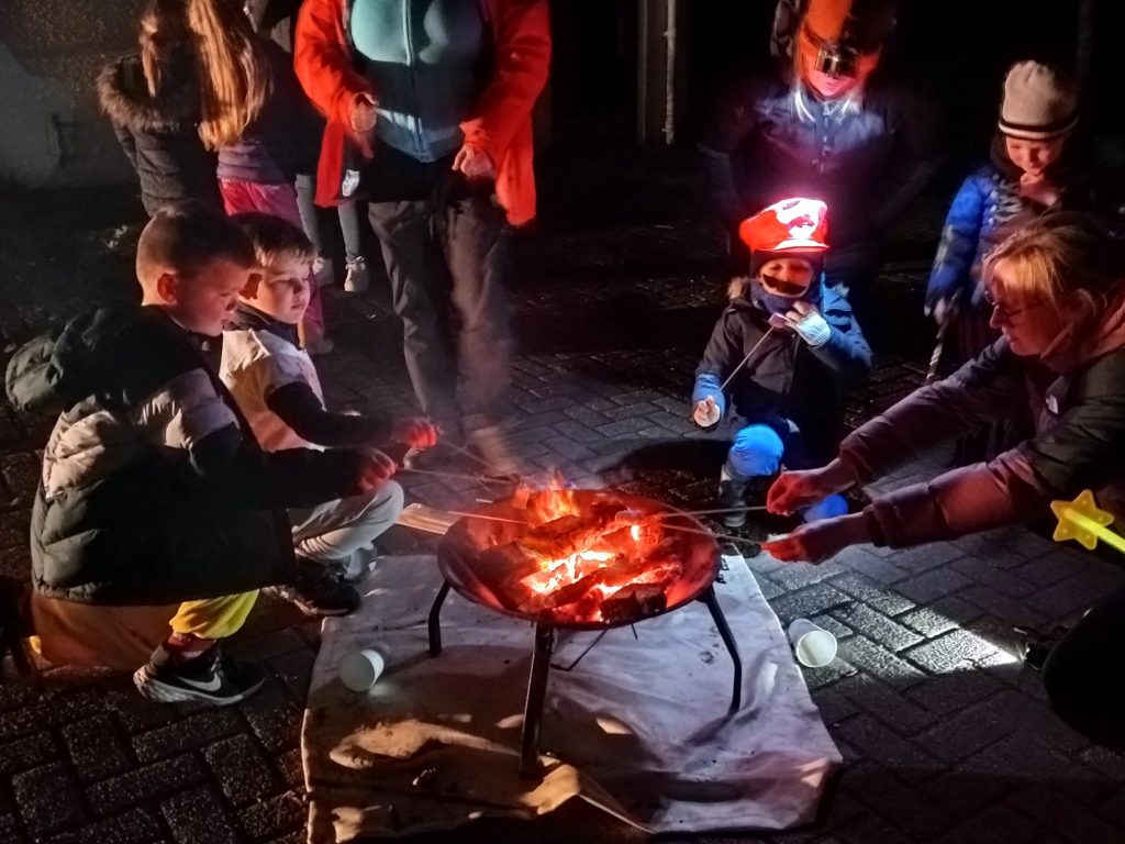 A group of adults and children are gathering around a campfire toasting marshmallows. It is night and some people are wearing head torches.