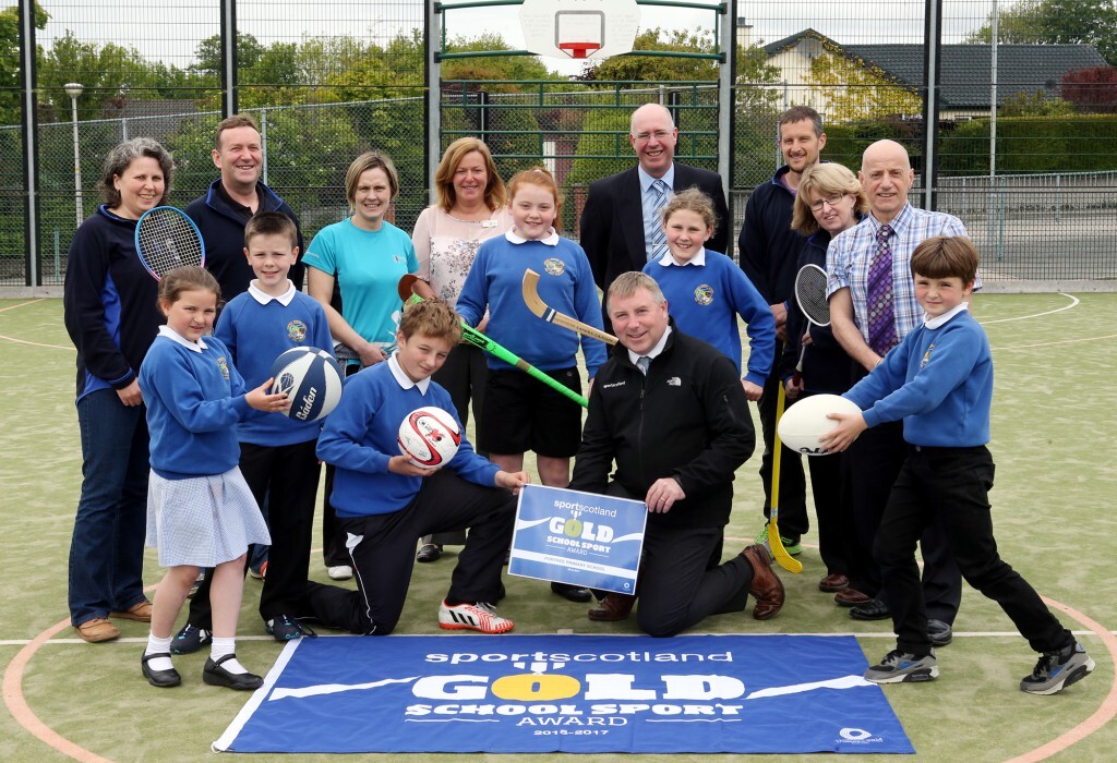 A group of adults and primary school pupils are on a sports pitch. The children are wearning blue school jumpers and holding a variety of sports equipment including rugby balls, footballs and shinty sticks. There are several adults with them including school staff, local councillors and representatives from sportscotland. On the ground in front of the group is a blue and gold flag.