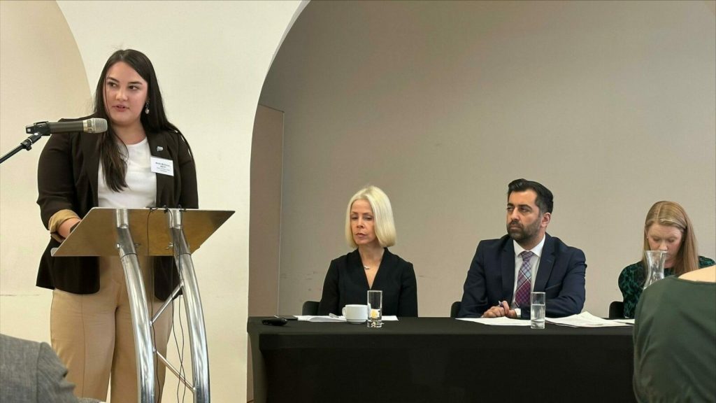 A young woman in smart clothes is standing at a podium giving a speech. Behind her is a table where three adults are sitting, listening to her.